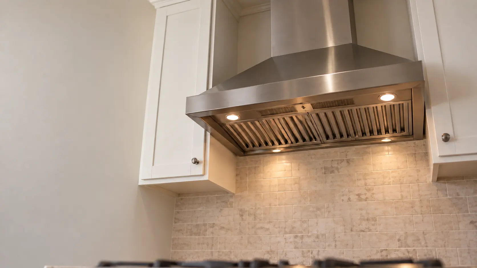 A kitchen with a stainless wall-mount range hood and white shaker cabinets, seen from below the range hood.