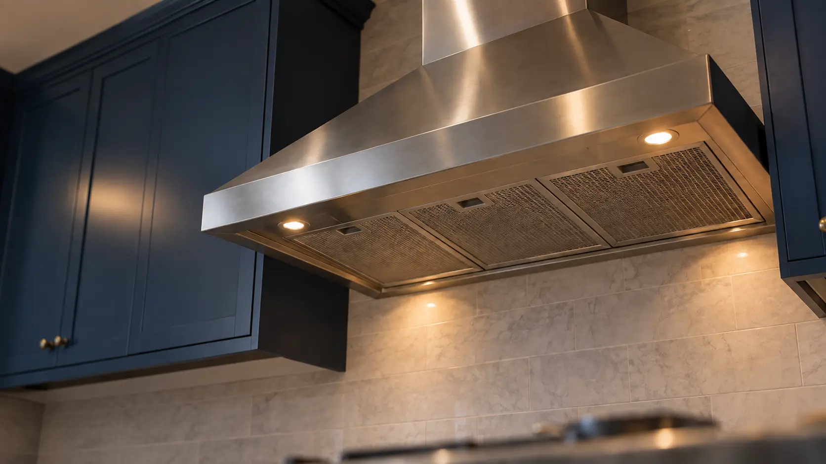 A kitchen with a stainless wall-mount range hood and navy painted cabinets, seen from below the range hood.