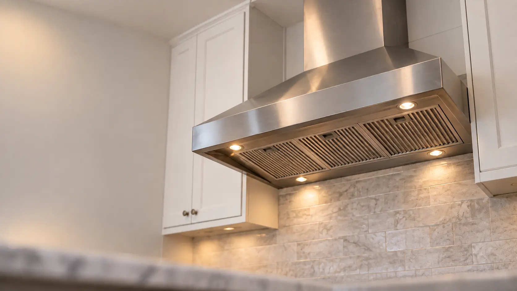 A kitchen with a stainless wall-mount range hood and white shaker cabinets, seen from below the range hood.