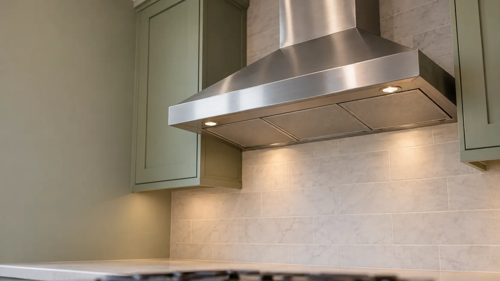 A kitchen with a stainless wall-mount range hood and sage green cabinets, seen from below the range hood.
