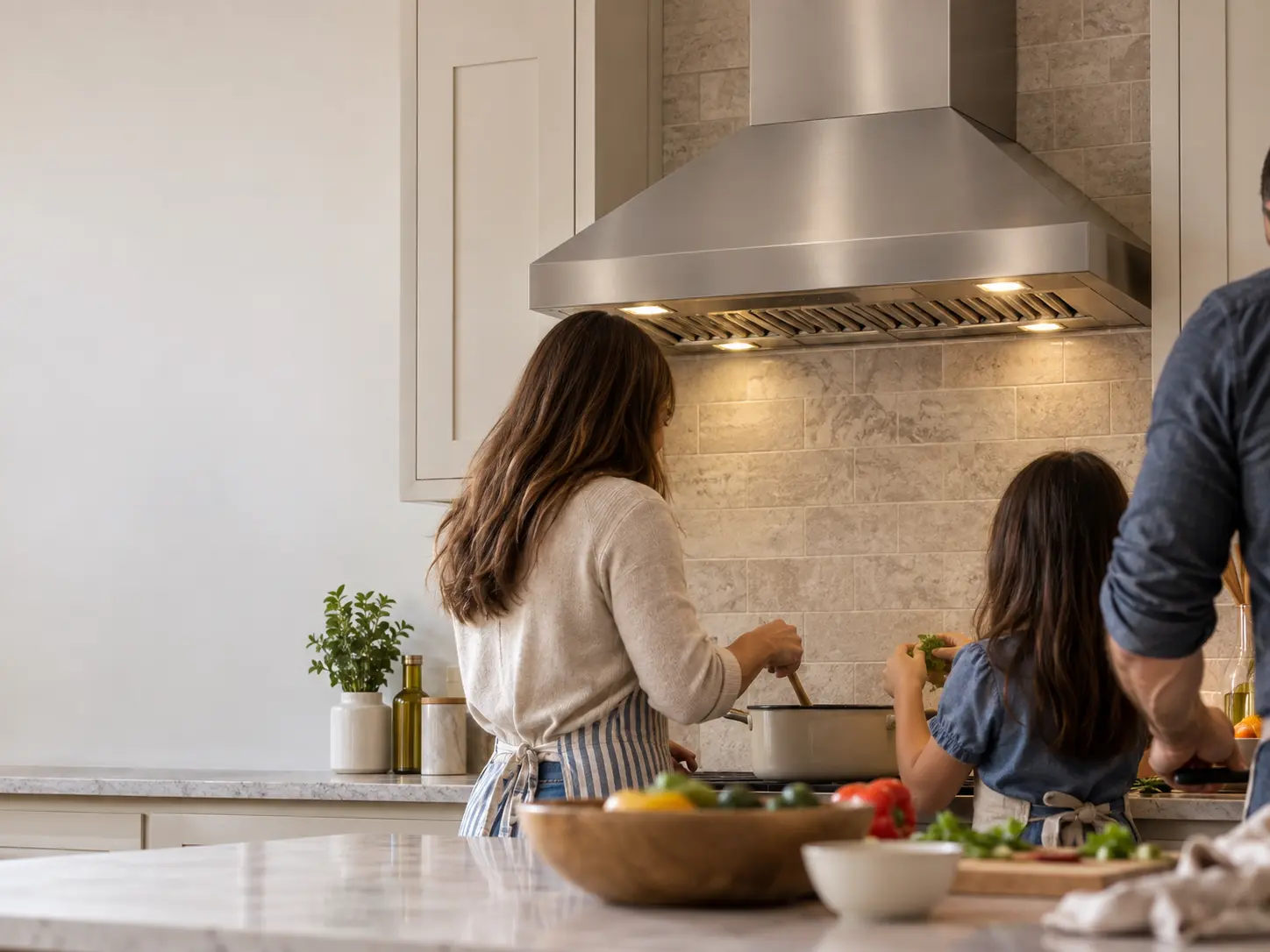 A family cooking together under a stainless range hood in a bright kitchen.