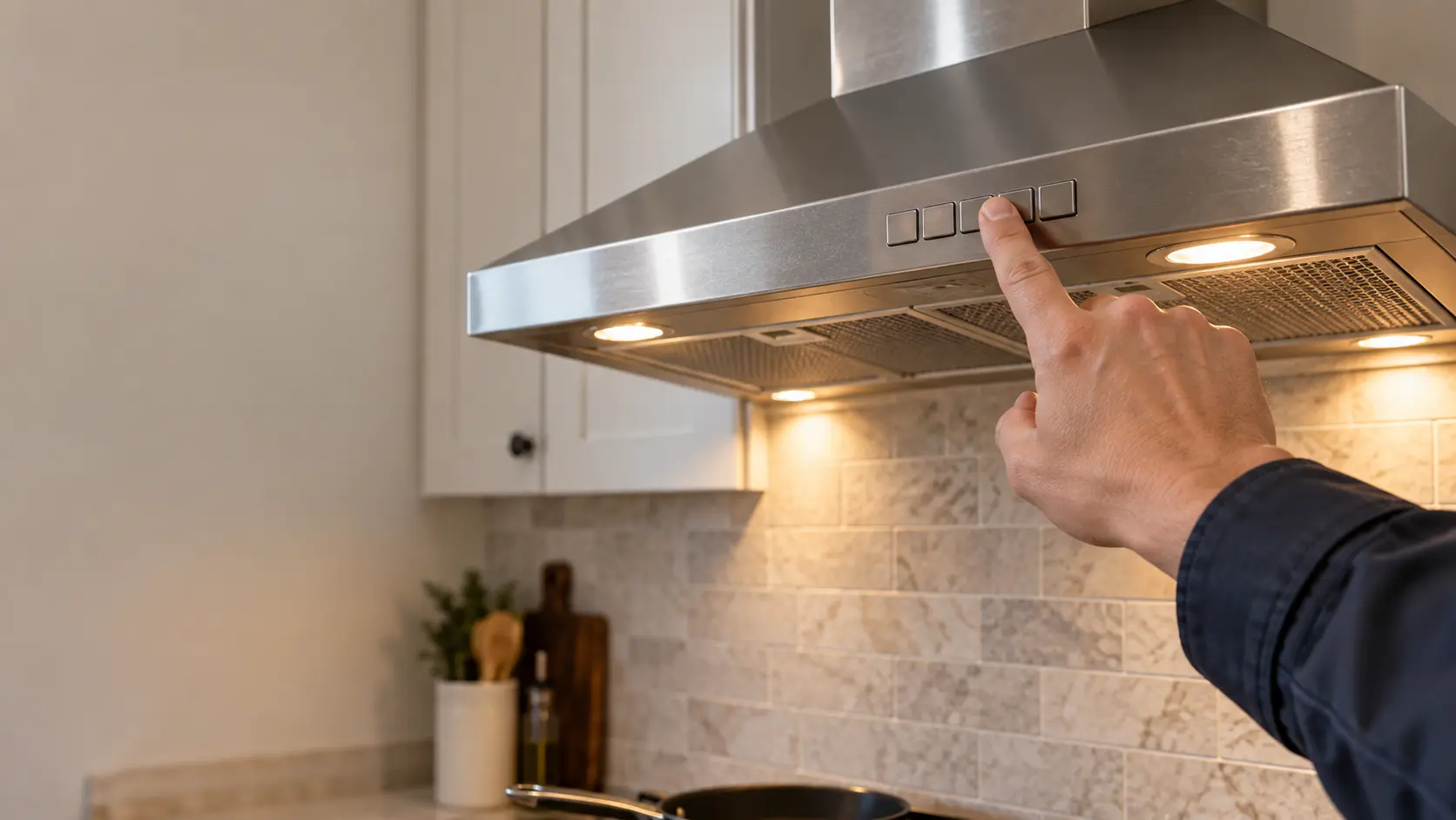 A close view of the front edge of a stainless under-cabinet range hood in a US home kitchen, its row of push keys set in the range hood's lip, a technician's clean fingertip pressing one blank key, the lamps beside it glowing, the cooking surface below soft and out of focus.