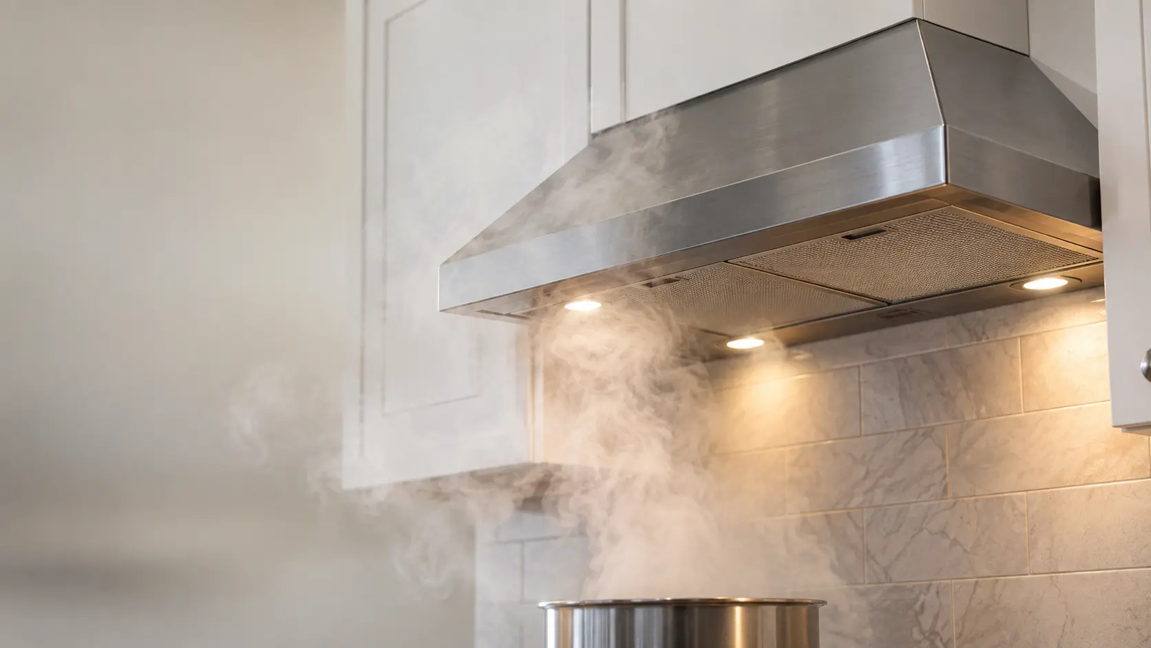 A stainless under-cabinet range hood below white kitchen cabinets fills the upper half of the frame, its lights on but steam from a simmering pot below curling out past the front edge of the range hood and up against the cabinet doors instead of into the canopy.