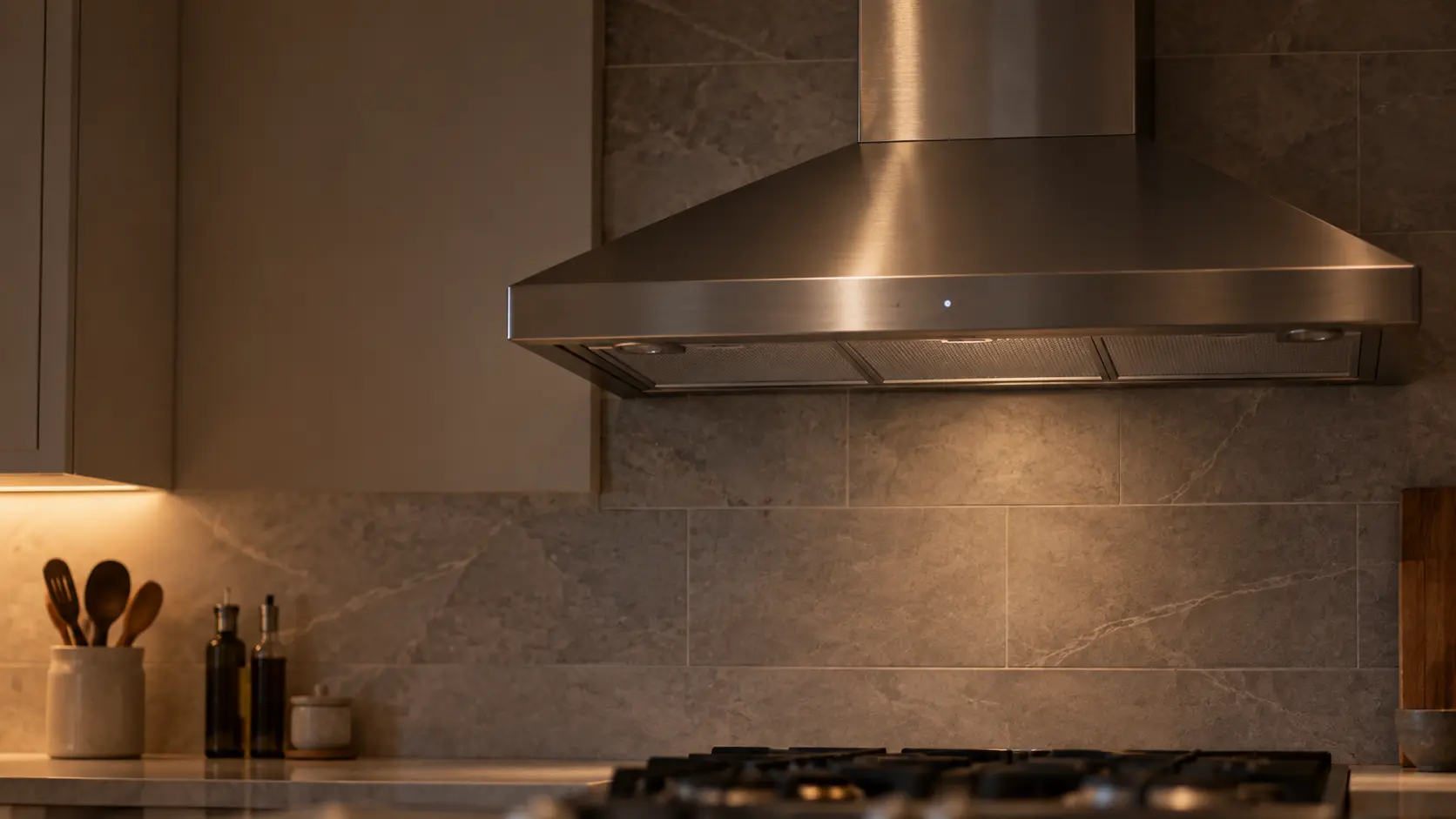 A stainless wall-mount chimney range hood on a tiled wall in a quiet, tidy kitchen late at night fills the upper half of the frame, its lights off but a small glowing indicator on the range hood's control strip, the burners below off and bare.