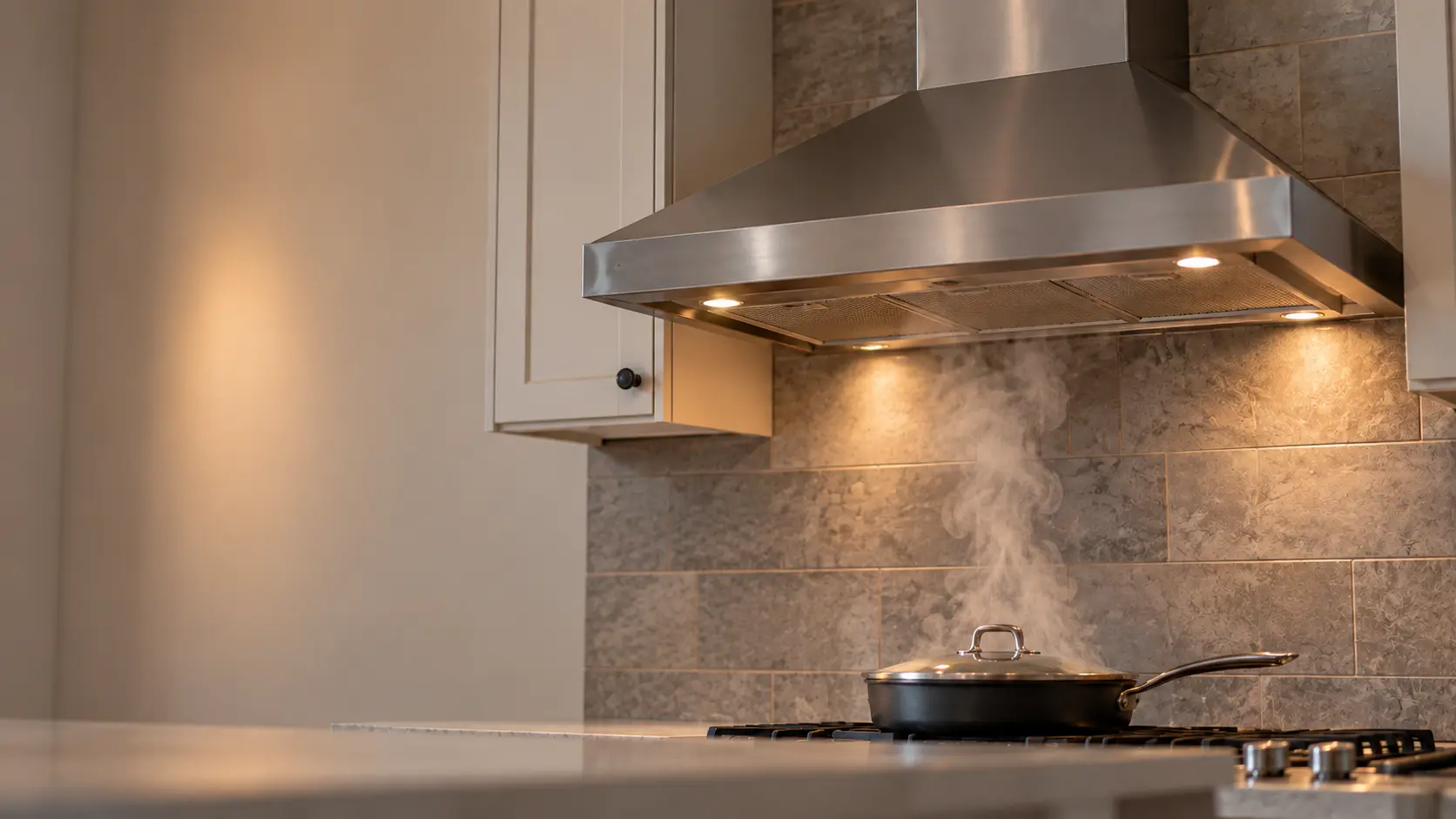 A stainless wall-mount chimney range hood on a tiled kitchen wall fills the upper half of the frame, dark gray smoke curling up into the range hood from a covered frying pan on the burner below, the pan's lid in place and the burner knob turned off.