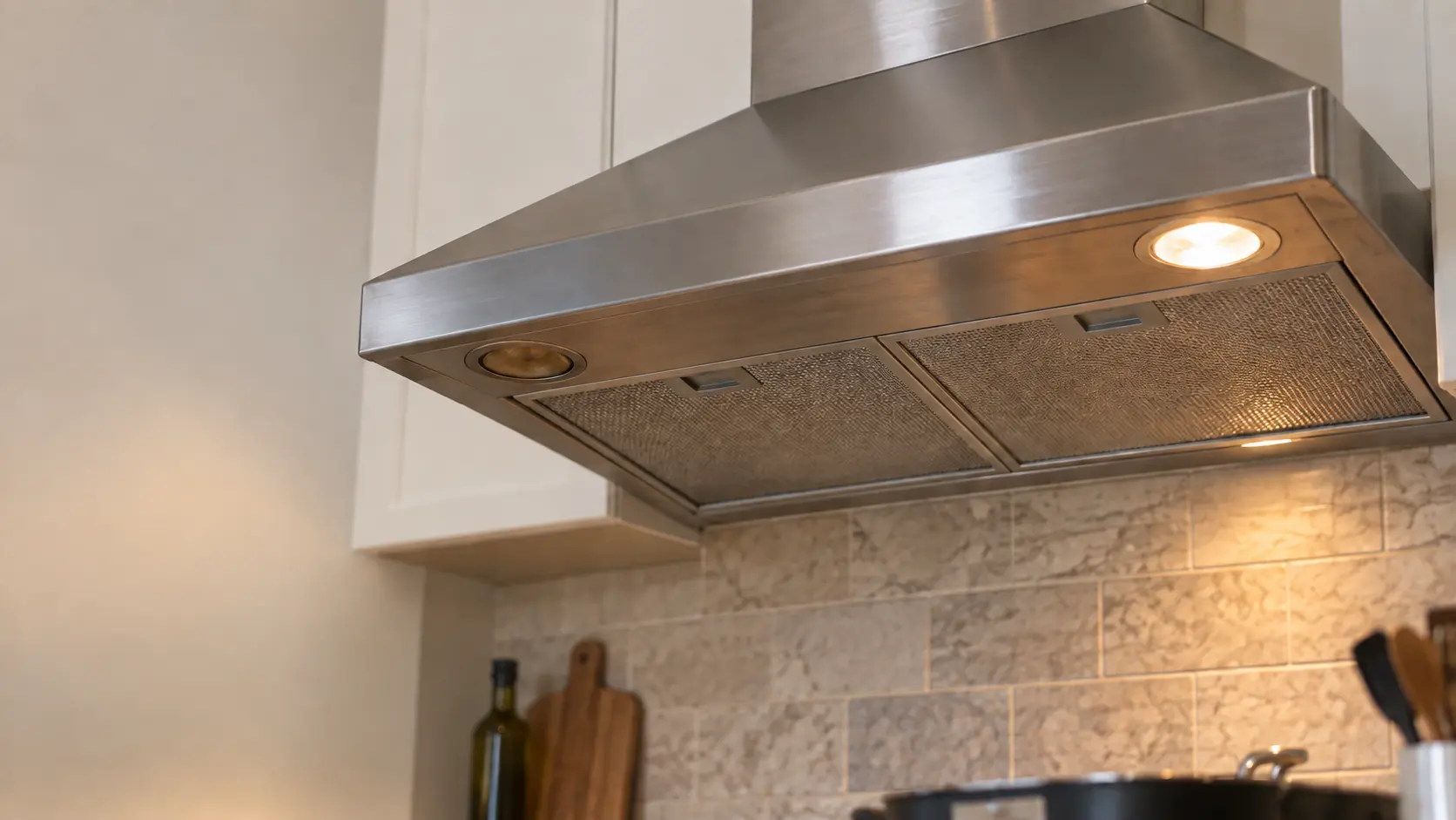 Close view looking up at the underside of a stainless under-cabinet range hood below white cabinets, two recessed lights in the range hood's underside, one glowing warm and the other dark with a slightly browned lens, the mesh filters between them clean.
