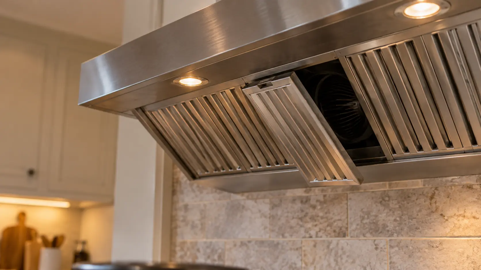 Close view from below of the underside of a stainless wall-mount range hood, one baffle filter hanging slightly crooked out of its track and the blower's intake visible in shadow behind it, the rest of the range hood's filters seated flat.