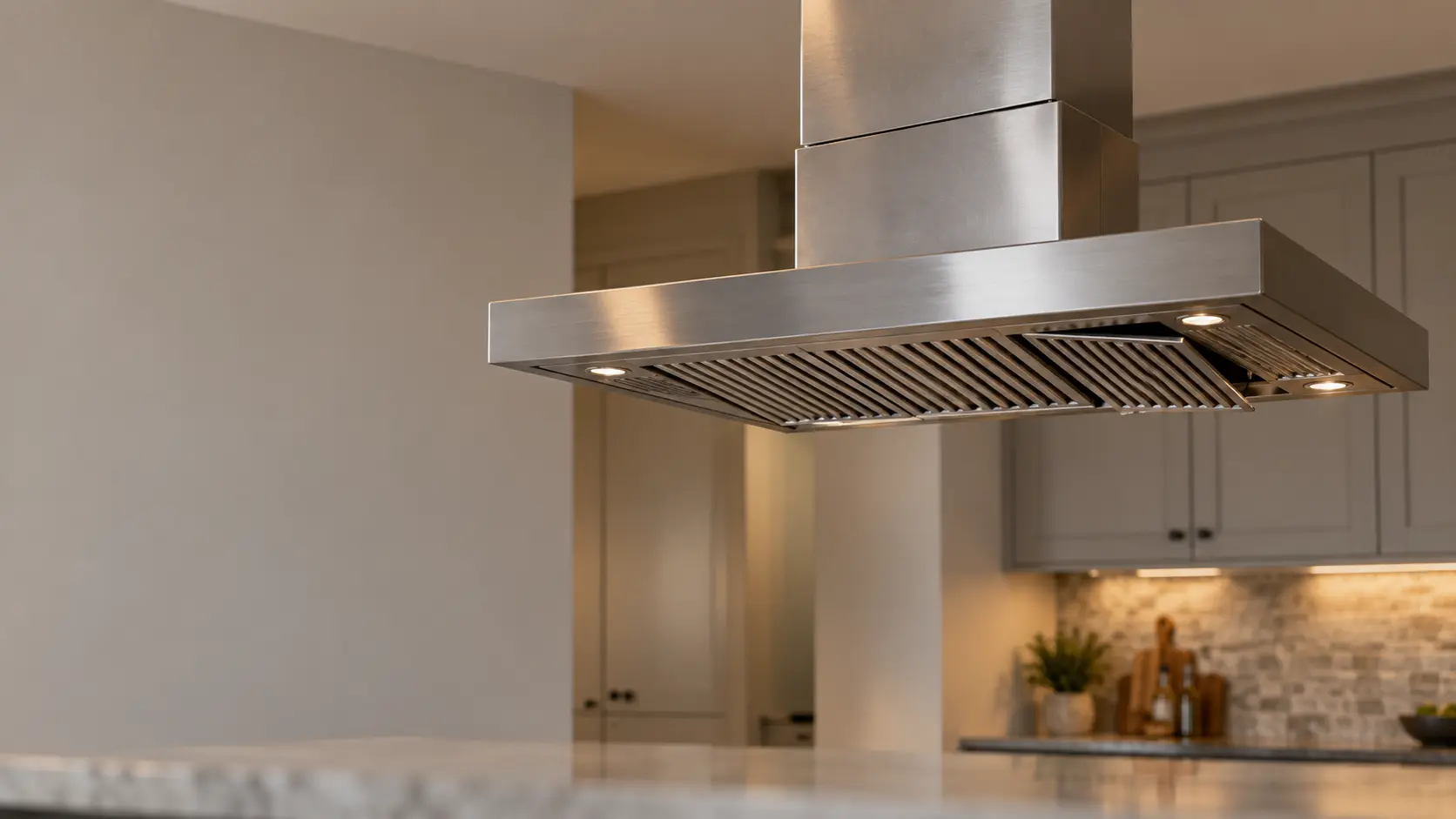 A stainless island range hood hanging from the ceiling over a kitchen island fills the upper half of the frame, its chimney cover sections slightly misaligned at the joint and one baffle filter visibly loose in its track on the range hood's underside, the island counter below empty and out of focus.