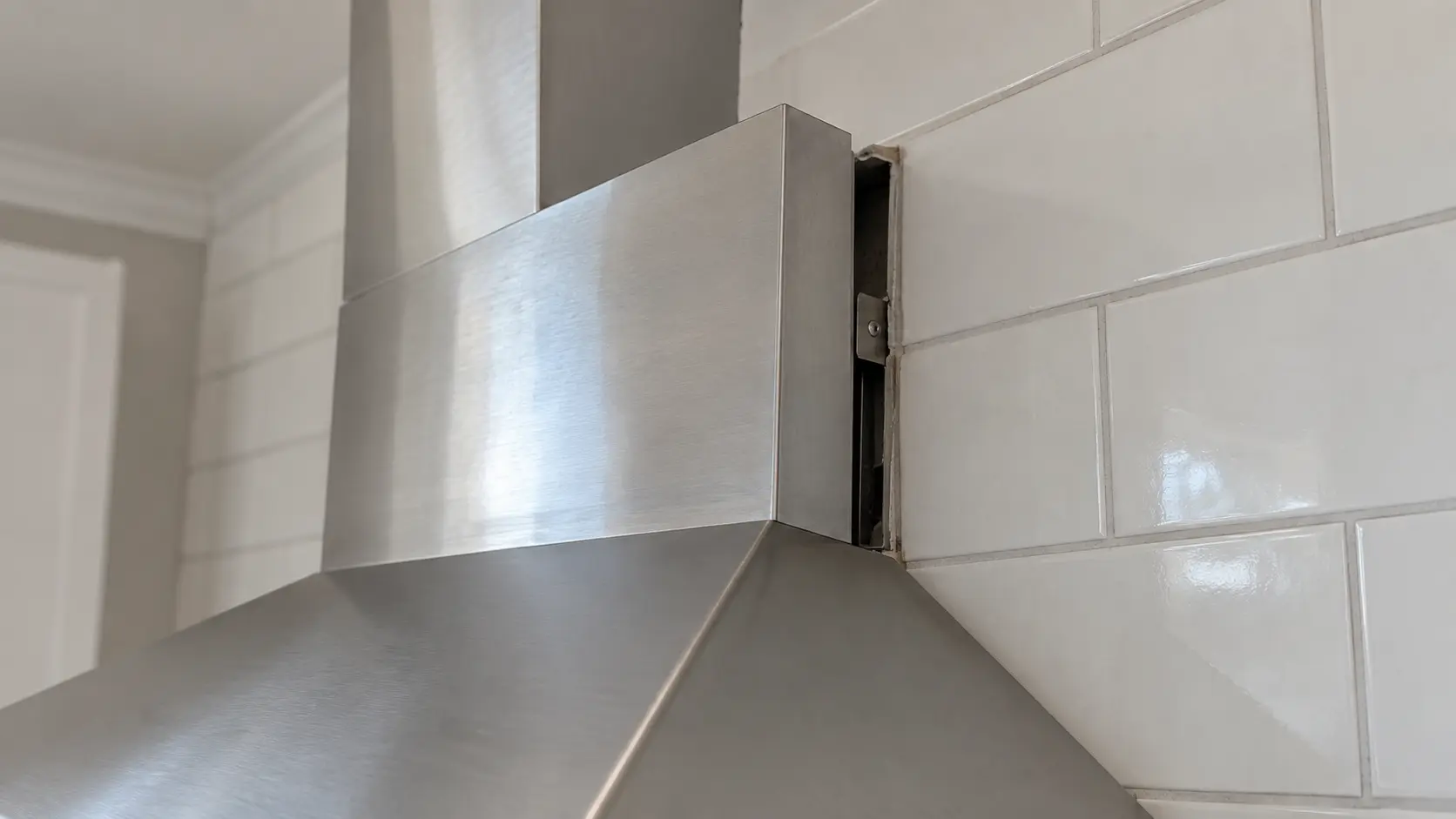 Close on a gap opening between the top of a chimney range hood and the tiled wall.