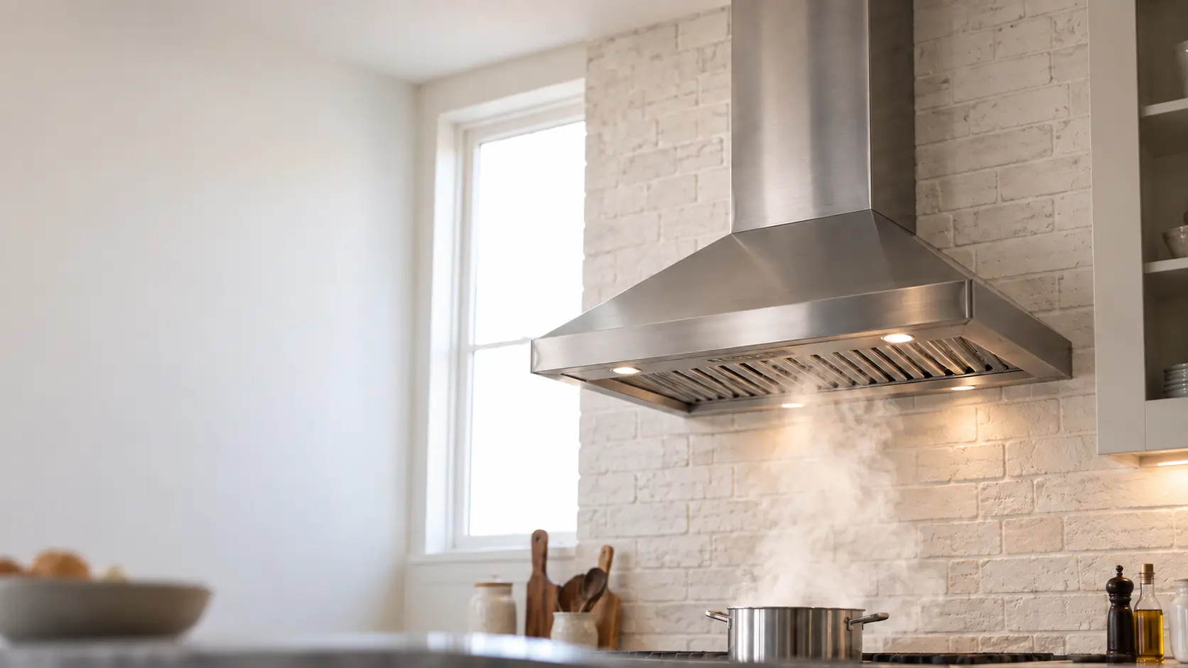 A brushed stainless wall-mount chimney range hood on a white brick kitchen wall, its chimney cover rising to the ceiling where the duct runs out of sight, steam rising from a pot on the burners into the range hood, the cooking surface soft and cropped at the bottom, a window beside it bright with daylight.