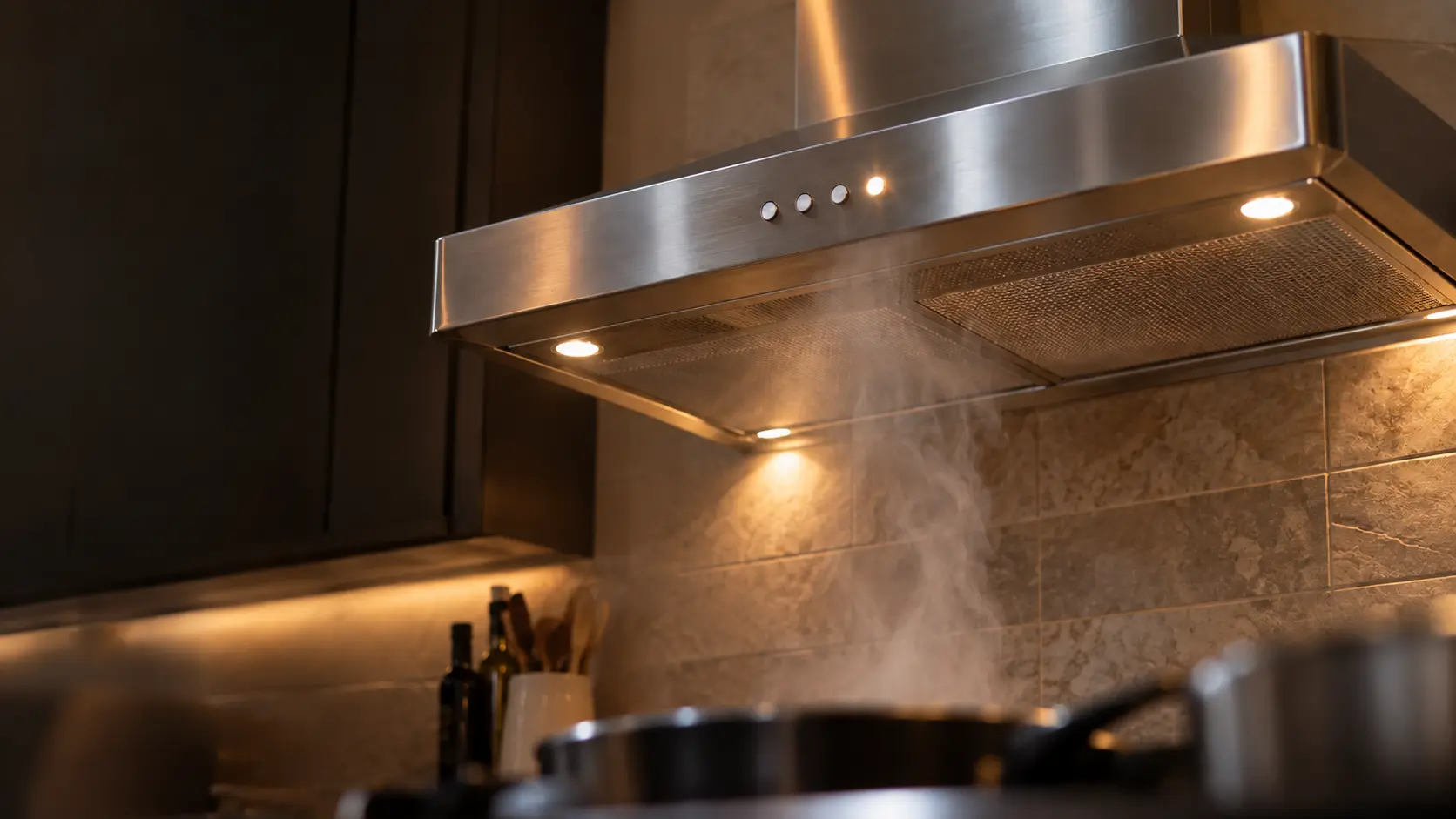 A close view from below of a stainless chimney range hood's front edge with its row of softly glowing keys, one key blinking, a thin plume of steam rising past it into the range hood, the burners below blurred and cropped, numbers.