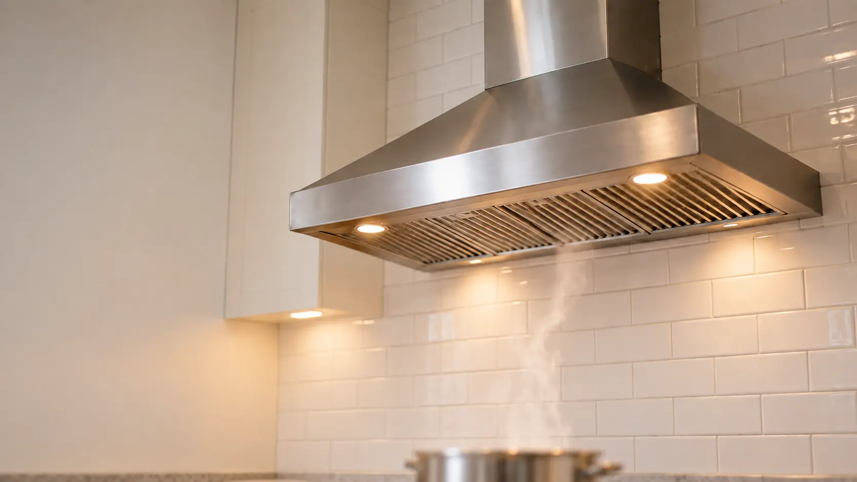 A wall-mount stainless chimney range hood on a white subway-tile wall, seen slightly from below with its filters in place and its lights on, a thin column of steam rising into the range hood from a pot on the burners cropped out of focus at the bottom edge.