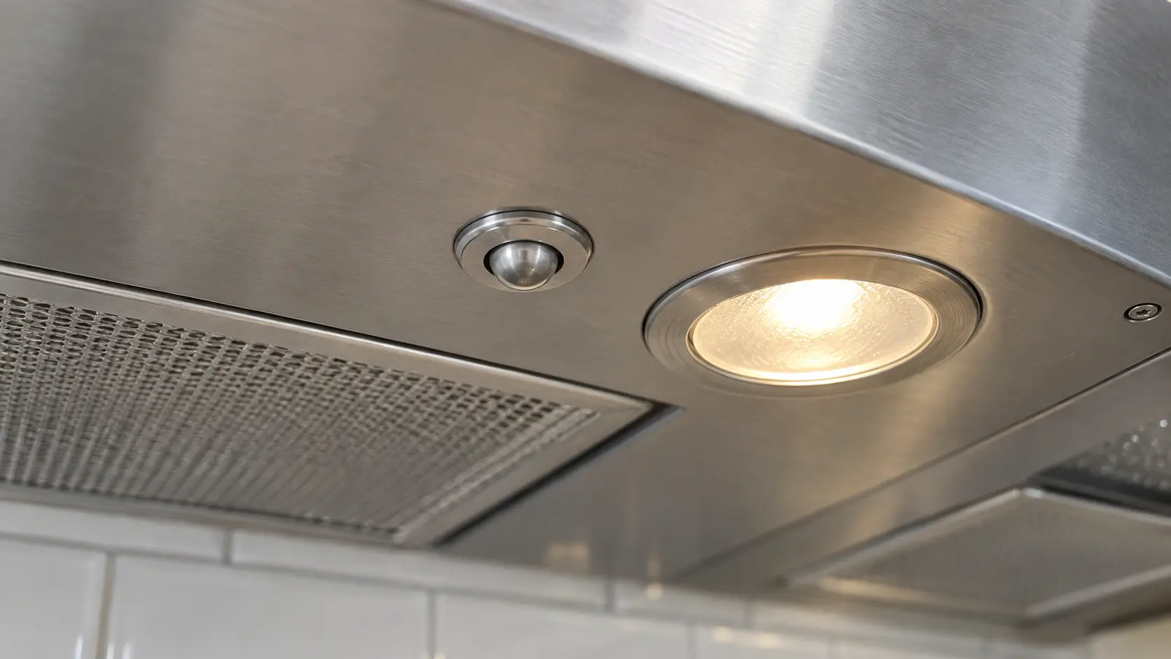 Close on the small round heat sensor on the underside of a range hood canopy beside its light.